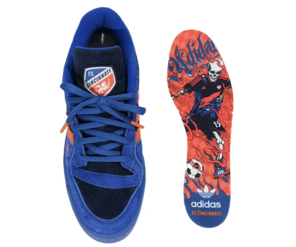 adidas Forum Low “All For Cincy” Blue Kit edition featuring the FC Cincinnati crest on the tongue, blue and orange suede upper with matching laces, and a custom insole illustrated with a fiery skeleton soccer player wearing the club’s kit, surrounded by smoke graphics and adidas FC Cincinnati branding at the heel.