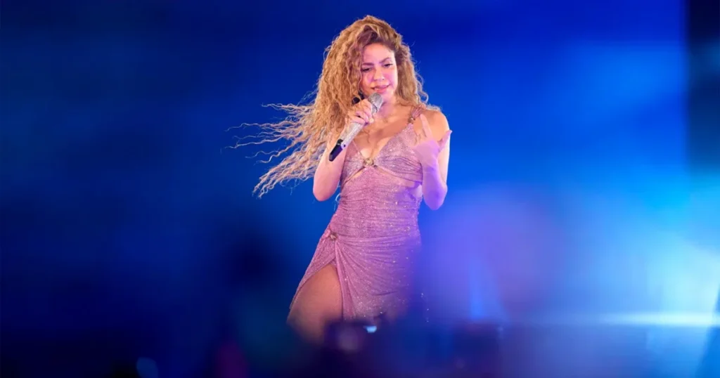 Shakira performs live on stage wearing a sparkling mauve dress with flowing curly hair, holding a silver microphone against a vibrant blue-lit background during her Spotify Anniversary celebration concert.