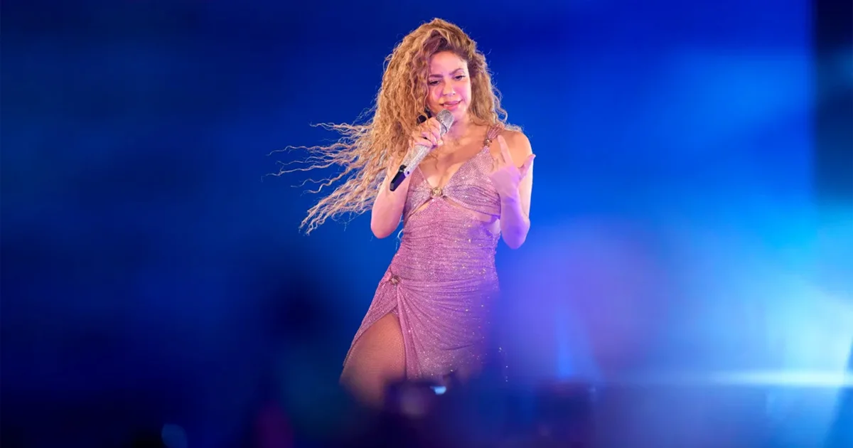 Shakira performs live on stage wearing a sparkling mauve dress with flowing curly hair, holding a silver microphone against a vibrant blue-lit background during her Spotify Anniversary celebration concert.
