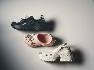 Flat lay of the Simone Rocha x Crocs Spring/Summer 2026 collection featuring three styles: the black Trailbreak sneaker adorned with crystals, the pink Classic Platform Clog decorated with oversized pearls and gems, and the ivory Ballerina Platform embossed with bow motifs and finished with metallic detailing — embodying Simone Rocha’s romantic yet utilitarian design language.