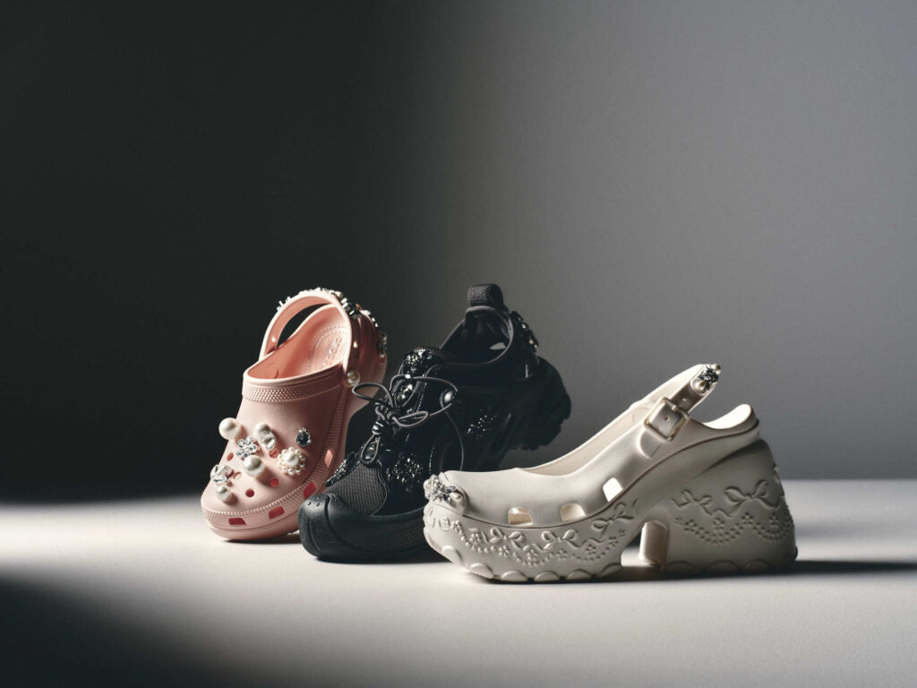 Studio image showcasing the Simone Rocha x Crocs Spring/Summer 2026 collection. From left to right: the embellished Classic Platform Clog in Pink Rose adorned with pearls and crystals, the Trailbreak sneaker in Black featuring rugged lace detailing, and the new Ballerina Platform in Oyster with embossed bow motifs and a sculpted heel — blending Crocs’ utility with Simone Rocha’s romantic elegance.