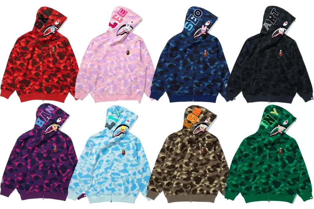 The complete lineup of BAPE × Stray Kids Shark Hoodies displayed in eight distinct camouflage colorways—red, pink, navy, black, purple, sky blue, brown, and green—each featuring embroidered initials and SKZOO character patches representing individual Stray Kids members. The design highlights BAPE’s signature shark-mouth hood combined with personalized details chosen by the idols.