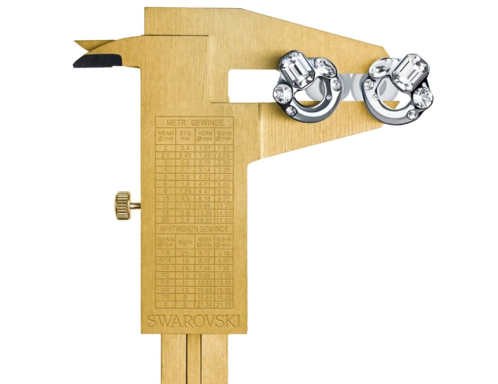 A gold caliper engraved with measurement markings and the Swarovski logo, used as a display stand holding a pair of Loop x Swarovski Experience 2 Limited Edition earplugs. The earplugs, encrusted with precision-cut Swarovski crystals, contrast against the matte gold instrument—symbolizing the collaboration’s blend of technical precision and luxurious craftsmanship.
