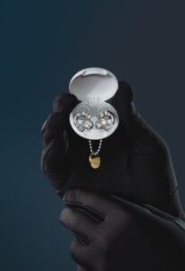 A pair of Loop x Swarovski Experience 2 Limited Edition earplugs, shimmering with precision-cut Swarovski crystals, displayed inside a glossy white circular case. The case is held delicately by gloved black hands against a deep navy background, with a small gold Loop tag hanging from a metallic chain beneath—highlighting the fusion of luxury design and acoustic innovation.