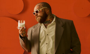 Teddy Swims stands against a burnt-orange textured backdrop, holding a glass of milk while wearing oversized white sunglasses, a taupe suit jacket, and an open-collar cream shirt. His tattooed neck and hands add a signature touch to his soulful, vintage-inspired look.