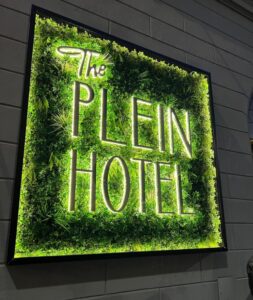 Illuminated exterior sign for The Plein Hotel in Milan, featuring glowing white letters surrounded by lush green foliage and mounted on a stone wall at night.
