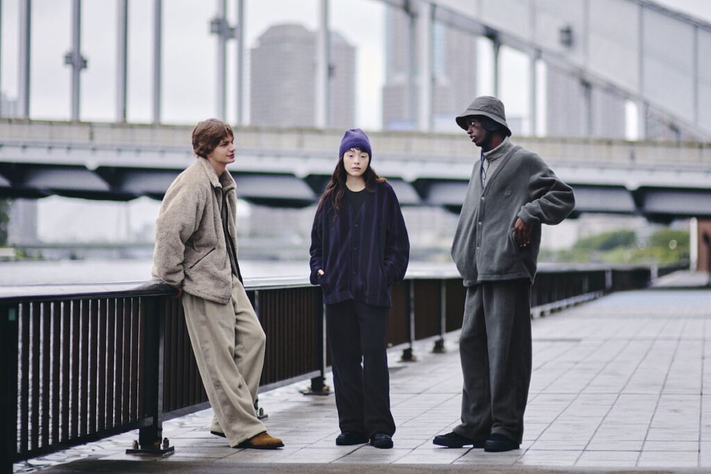 Three models wearing pieces from the UNIQLO × NEEDLES Fleece Collection stand by a riverside walkway in Tokyo. Each wears oversized, minimalist fleece garments in neutral tones—beige, charcoal, and deep purple—styled with relaxed wide-leg pants and understated accessories. The scene captures a modern, genderless aesthetic that blends Japanese craftsmanship with effortless streetwear comfort.