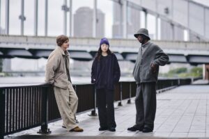 Three models wearing pieces from the UNIQLO × NEEDLES Fleece Collection stand by a riverside walkway in Tokyo. Each wears oversized, minimalist fleece garments in neutral tones—beige, charcoal, and deep purple—styled with relaxed wide-leg pants and understated accessories. The scene captures a modern, genderless aesthetic that blends Japanese craftsmanship with effortless streetwear comfort.