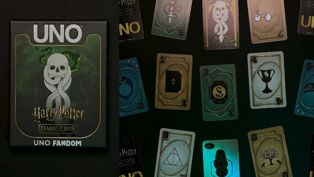 Mattel Creations’ UNO: Harry Potter Dark Arts deck displayed under dim light. On the left, the black box features a glowing green skull and serpent emblem representing the Dark Mark. On the right, the laid-out cards reveal intricate gothic artwork inspired by Horcruxes, Hogwarts houses, and magical relics, each with metallic foiling and a cursed, vintage finish.