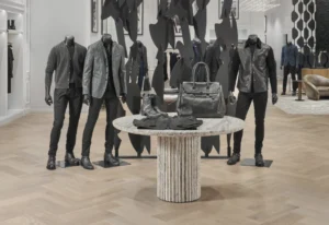 The space has light wood herringbone flooring, sculptural black metal partitions, and modern lounge seating in the background, embodying the brand’s refined rock aesthetic.