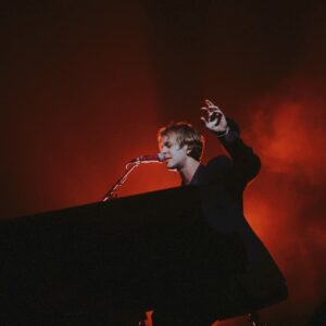 Tom Odell performs at the piano under a dramatic red spotlight, his hand raised expressively as he sings, capturing the emotional depth of “When I Close My Eyes."