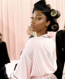 Angel Reese prepares backstage at the Victoria’s Secret Fashion Show 2025 wearing the brand’s signature pink-and-white striped robe with crystal-studded wings on the back, her hair set in large rollers and makeup glowing under soft pink lighting.