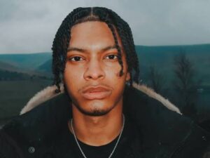 Portrait of Nemzzz, the Manchester-born rapper, standing outdoors against a moody hillside backdrop. He wears a dark jacket with fur trim, his expression calm and focused — reflecting the introspective tone of his music.