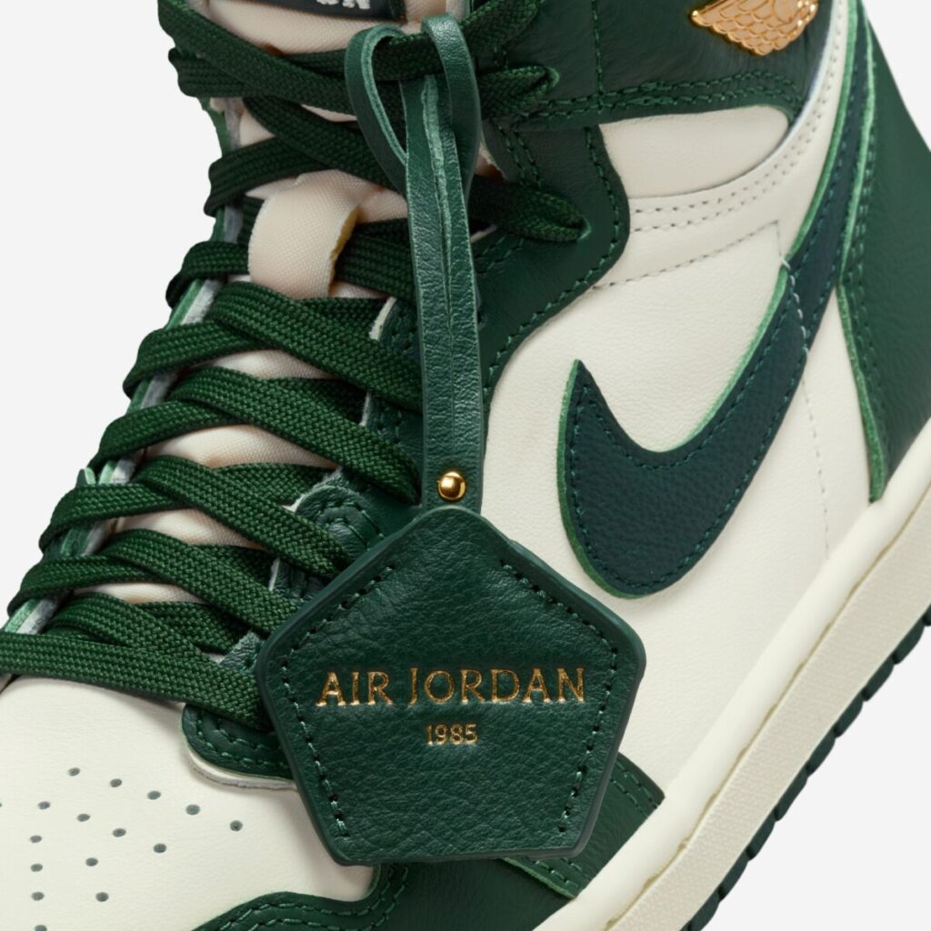 Close-up of the Air Jordan 1 High OG WMNS “Fir,” highlighting the green leather hang tag embossed with “Air Jordan 1985” in gold, dark green laces, smooth ivory leather upper, and gold Wings logo near the collar.
