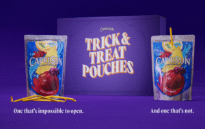 Halloween-themed promotional image for Capri Sun. Two juice pouches are shown side by side against a deep purple background. The pouch on the left has several bent, broken yellow straws lying in front of it, labeled “One that’s impossible to open.” The pouch on the right has an inserted straw, labeled “And one that’s not.” Behind them, a purple Capri Sun box reads “Trick & Treat Pouches” in bold golden letters with spiderweb graphics, highlighting the humor of the campaign