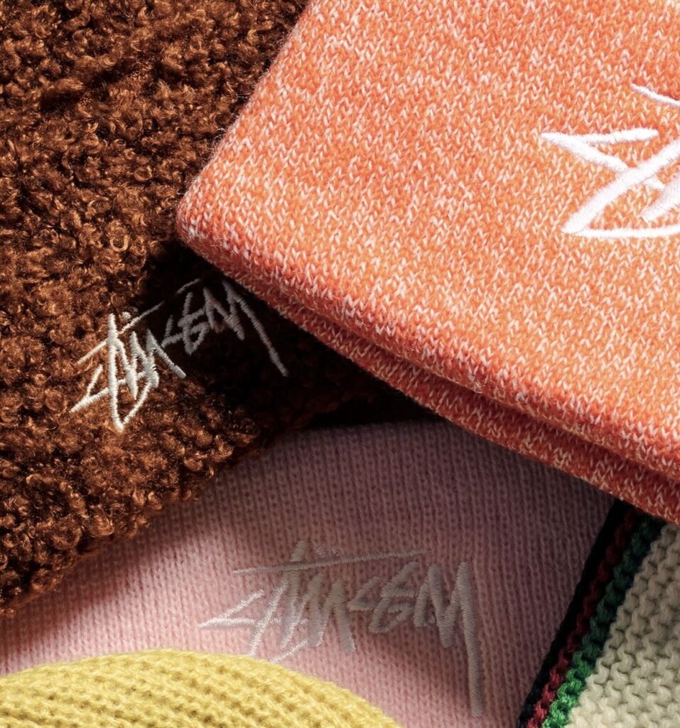 Close-up photograph of Stüssy Holiday 2025 Collection beanies in warm seasonal tones—brown boucle, orange marled knit, pastel pink rib, mustard yellow wool, and cream with green-red stripe—each embroidered with the iconic Stüssy script logo, highlighting texture and color diversity.
