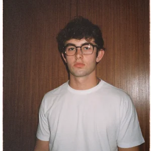 Ian, wearing round glasses and a plain white T-shirt, standing against a wooden-paneled background with a calm, neutral expression.