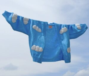 A bright blue cardigan with fluffy white cloud patterns, photographed against a matching blue sky backdrop, creating the illusion that the sweater is floating among the clouds.
