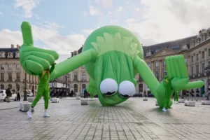 Installation view of Alex Da Corte’s Kermit the Frog, Even (2025) at Place Vendôme, Paris — a monumental inflatable sculpture of Kermit slumped forward with outstretched arms, his oversized eyes gazing downward, while performers in matching green suits interact playfully around the fallen figure against the square’s historic architecture.