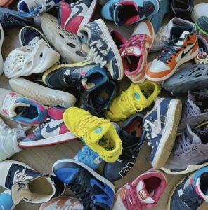 A colorful pile of collectible shoes, including Nike Dunks, Air Jordans, Yeezy Foam Runners, and bright yellow Grateful Dead SB Dunk Lows, scattered on a wooden floor, showcasing the diversity and vibrancy of footwear culture.