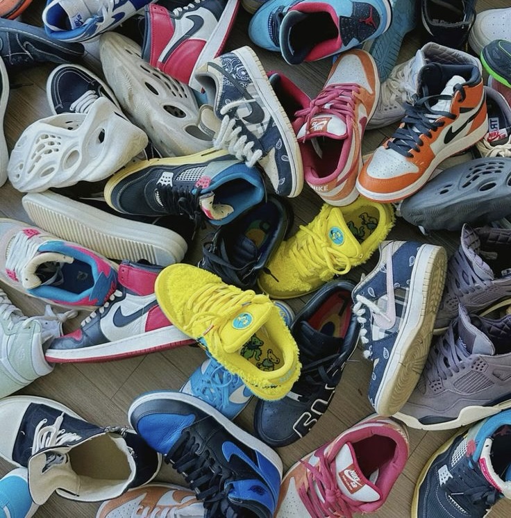 A colorful pile of collectible shoes, including Nike Dunks, Air Jordans, Yeezy Foam Runners, and bright yellow Grateful Dead SB Dunk Lows, scattered on a wooden floor, showcasing the diversity and vibrancy of footwear culture.
