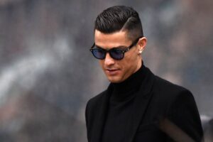 Cristiano Ronaldo wearing dark sunglasses and a black turtleneck with a blazer, looking down with a composed expression.