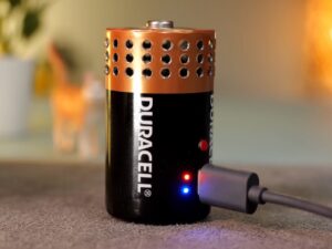 A DIY Bluetooth speaker disguised inside a Duracell battery casing, modified with precision-drilled sound holes, indicator lights, and a charging port, placed on a table next to a smartphone playing music.