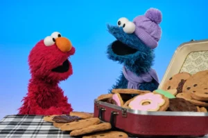 Elmo and Cookie Monster from Sesame Street sit at a checkered table with an open suitcase full of cookies and pastries. Cookie Monster wears a purple winter hat and scarf, while Elmo looks on excitedly against a bright blue background, capturing their playful spirit ahead of the 2026 Winter Olympics.