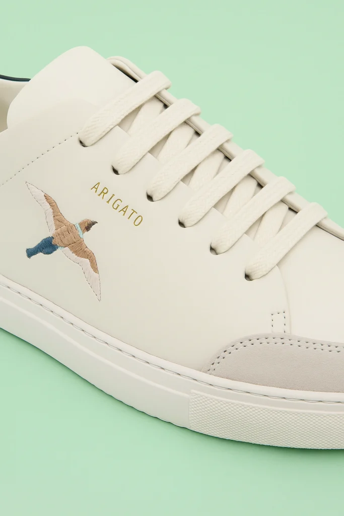 Close-up of the Axel Arigato Clean 90 Bee Bird sneaker in off-white leather, featuring gold “ARIGATO” branding and a detailed multicolour embroidered bird motif, highlighting fine stitching and premium craftsmanship against a light green background.