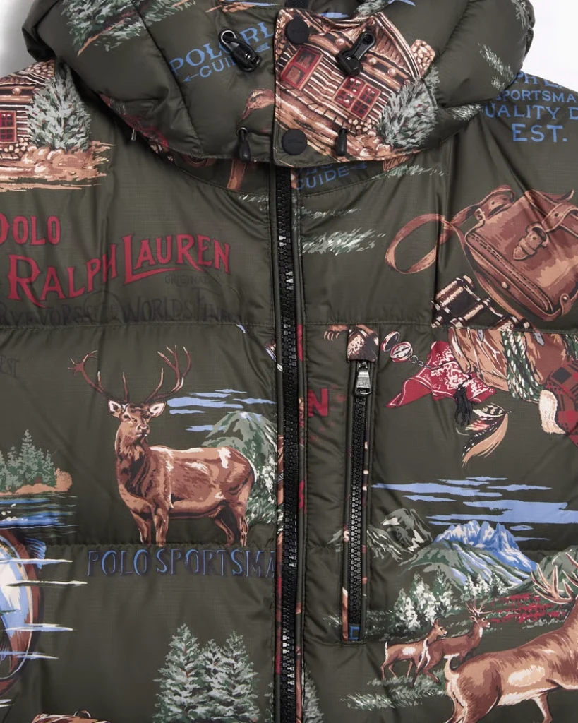 Close-up detail of the Polo Ralph Lauren Gorham Sportsman Ripstop Graphic Down Jacket in olive green. The image highlights the printed outdoor-inspired artwork featuring a stag, mountain scenery, log cabins, and vintage travel motifs alongside the red Polo Ralph Lauren logo. The ripstop texture, zip closure, and adjustable hood emphasize its rugged construction and heritage sportsman aesthetic.