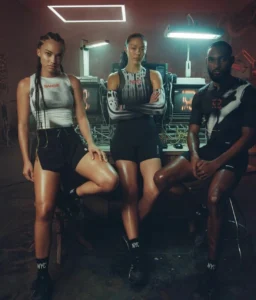 Three models pose in a dimly lit industrial studio surrounded by computer equipment, wearing Bandit athletic apparel from the NYC-themed collection — sleek black shorts, gradient performance tops, and NYC-branded socks under fluorescent lights.