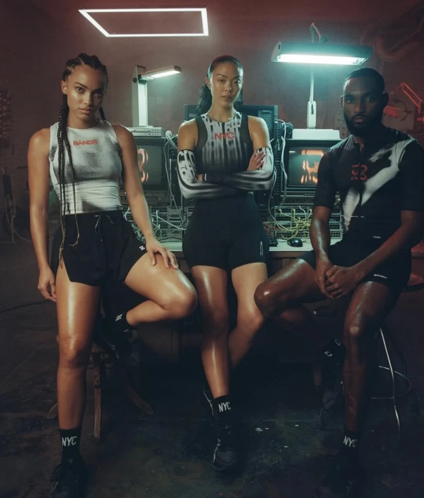 Three models pose in a dimly lit industrial studio surrounded by computer equipment, wearing Bandit athletic apparel from the NYC-themed collection — sleek black shorts, gradient performance tops, and NYC-branded socks under fluorescent lights.