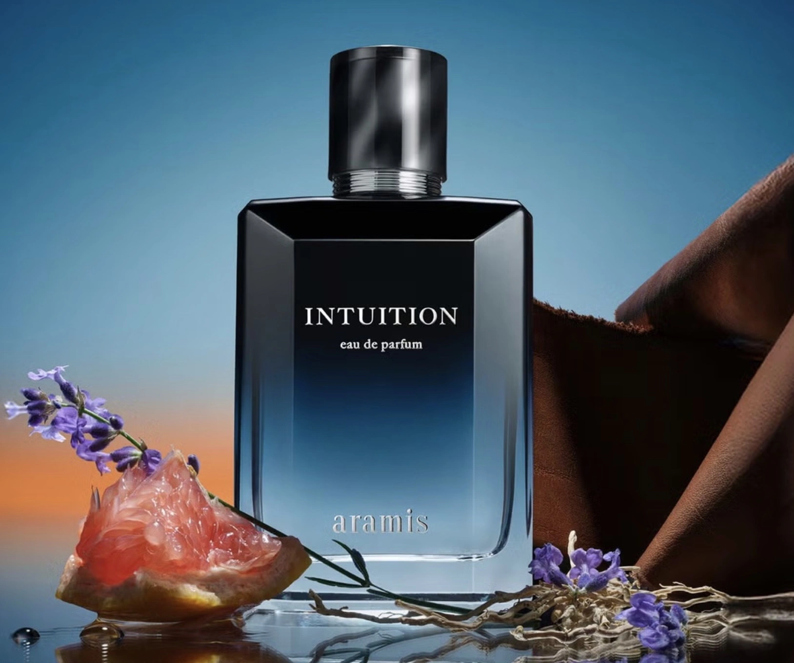 A close-up studio image of Intuition Eau de Parfum by Aramis. The square glass bottle features a gradient design shifting from deep midnight blue at the top to clear at the base, with “INTUITION” and “aramis” printed in silver text. It’s set against a softly blurred sky background of orange and blue hues, surrounded by fresh lavender sprigs, a grapefruit wedge, and a fold of brown leather fabric — visually representing the fragrance’s key notes of citrus, lavender, and leather.