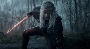 Geralt of Rivia, dressed in dark studded armor, crouches in a misty forest with a bloodied face and glowing steel sword drawn, ready for battle in a tense scene from The Witcher.