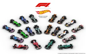 Overhead view of the complete 2025 Formula 1 grid recreated in 1:64 scale by Hot Wheels, featuring ten officially licensed team cars arranged diagonally on a white surface. The miniature lineup includes the vibrant red Ferrari HP, green Aston Martin Aramco, orange McLaren, navy and yellow Red Bull, silver Mercedes, blue Williams, pink Alpine, white Haas, and other team liveries. The F1 and Hot Wheels logos appear above, marking the collection’s status as the official licensed Formula 1 die-cast series.