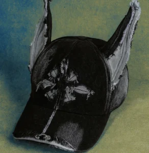 Front view of the GENZERO Doberman Hat in black washed-jersey fabric, featuring distressed stitching, torn cross-shaped detailing on the crown, and long frayed ears, photographed against a textured blue and gold background.