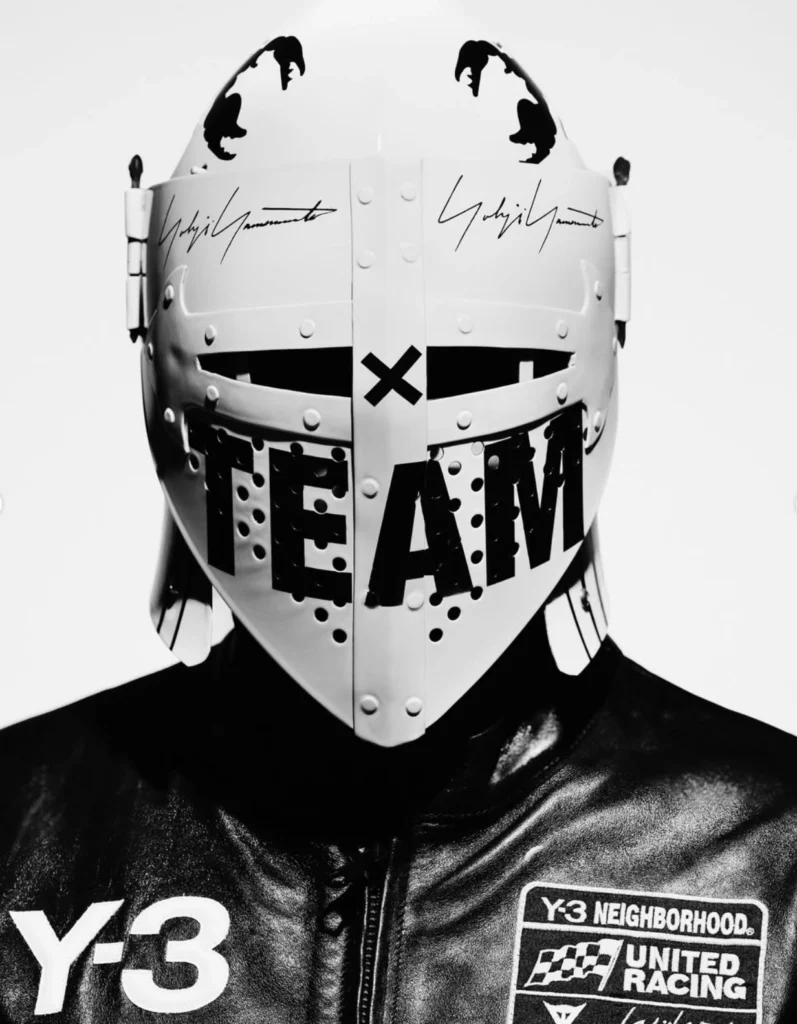 Black-and-white close-up of a model wearing a futuristic Y-3 × NEIGHBORHOOD Fall/Winter 2025 motorcycle helmet emblazoned with the word “TEAM” and Yohji Yamamoto’s signature, paired with a leather racing jacket featuring Y-3 and NEIGHBORHOOD United Racing logos.