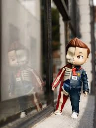 XXRAY PLUS: Buddy Lee collectible by Mighty Jaxx standing beside a reflective window. The half-anatomical vinyl figure reveals its skeletal interior on one side, while the other half displays Buddy Lee’s classic denim outfit and red scarf.