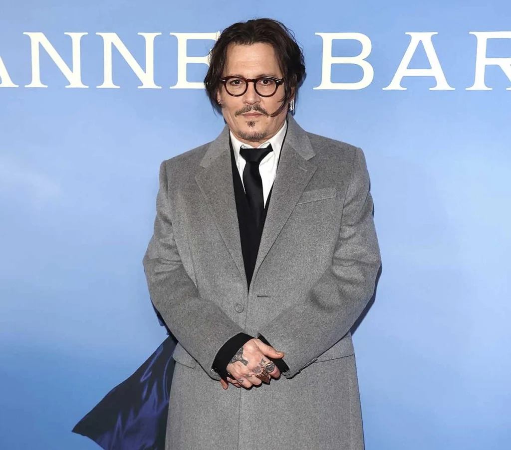 Johnny Depp stands against a light blue backdrop wearing a grey overcoat, black tie, and round glasses, with his hands clasped — a composed, vintage look that complements his forthcoming role in Ebenezer: A Christmas Carol.