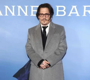 Johnny Depp stands against a light blue backdrop wearing a grey overcoat, black tie, and round glasses, with his hands clasped — a composed, vintage look that complements his forthcoming role in Ebenezer: A Christmas Carol.