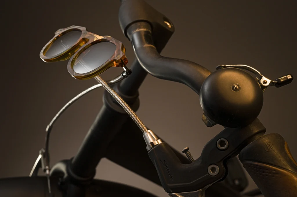 Vinylize CYKLO eyewear with amber bio-acetate frames and recycled bicycle brake cable temples rests on the handlebars of a matte black bicycle. The image highlights the connection between cycling mechanics and sustainable eyewear design central to the CYKLO collection.
