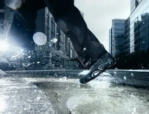 Dynamic action shot of a person running through a puddle in an urban setting, wearing the black PUMA x San San Gear Mostro “Caged” sneakers. Water splashes around as the shoe’s sculpted sole and futuristic details catch the light against a backdrop of glass high-rises and city reflections.