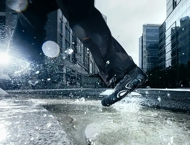 Dynamic action shot of a person running through a puddle in an urban setting, wearing the black PUMA x San San Gear Mostro “Caged” sneakers. Water splashes around as the shoe’s sculpted sole and futuristic details catch the light against a backdrop of glass high-rises and city reflections.