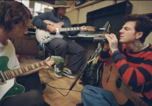 The Neighbourhood members in a recording studio session. One musician sings into a microphone while playing acoustic guitar, another plays an electric guitar, and a third sits in the background with a semi-hollow electric guitar. The setting is intimate, with warm lighting, casual clothing, and a relaxed creative atmosphere, capturing the band’s return to songwriting during their ultraSOUND era.