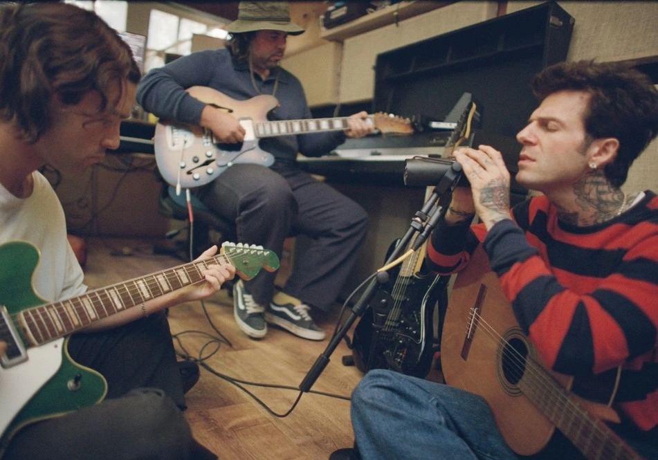 The Neighbourhood members in a recording studio session. One musician sings into a microphone while playing acoustic guitar, another plays an electric guitar, and a third sits in the background with a semi-hollow electric guitar. The setting is intimate, with warm lighting, casual clothing, and a relaxed creative atmosphere, capturing the band’s return to songwriting during their ultraSOUND era.