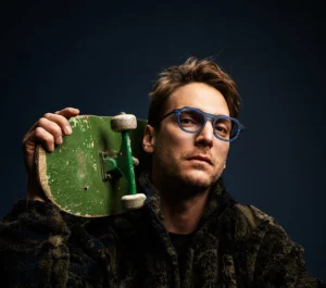 A man wearing blue Vinylize CYKLO eyewear poses against a dark background, holding a worn green skateboard on his shoulder. The portrait captures a blend of craftsmanship and urban attitude, reflecting the collection’s ethos of sustainability and individuality.