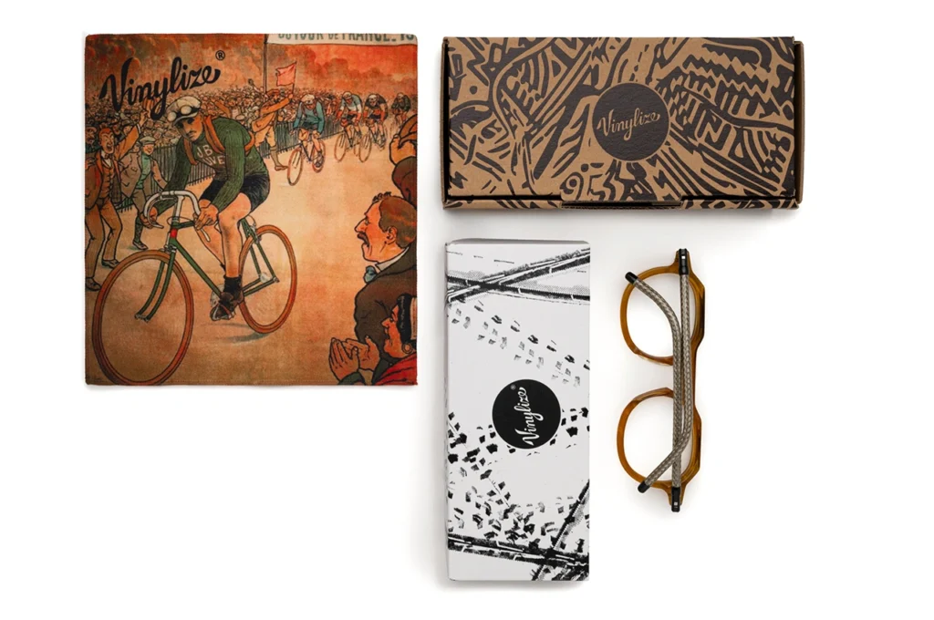 Vinylize CYKLO eyewear set displayed with packaging and accessories, including amber-framed glasses with recycled bicycle cable temples, a vintage cycling-themed cleaning cloth, and two custom-printed boxes featuring the Vinylize logo. The layout emphasizes the collection’s sustainable craftsmanship and cycling heritage.