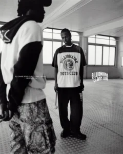 Black-and-white campaign image from Billionaire Boys Club Pre-Spring 2026, featuring two male models in a minimalist indoor setting. One wears a varsity-style short-sleeve jersey with the Billionaire Boys Club crest logo and relaxed trousers, while the other appears partially in frame wearing a hooded top and patterned cargo pants. The atmosphere evokes collegiate sports culture and Southern rhythm, emphasizing the brand’s blend of streetwear and athletic heritage.