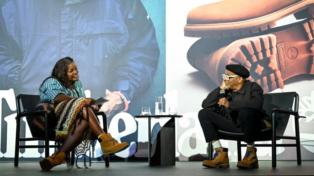 Spike Lee and a moderator share a conversation on stage during Timberland’s “Advice of an Icon” event. Both are seated against a large backdrop featuring Timberland’s signature boot sole and branding. The moderator smiles while holding notes, wearing a striped shirt, multicolored skirt, and yellow Timberland boots. Spike Lee, dressed in a black beret, dark jacket, and matching boots with blue laces, listens intently, microphone in hand, embodying the relaxed wisdom and creative spirit central to the campaign.