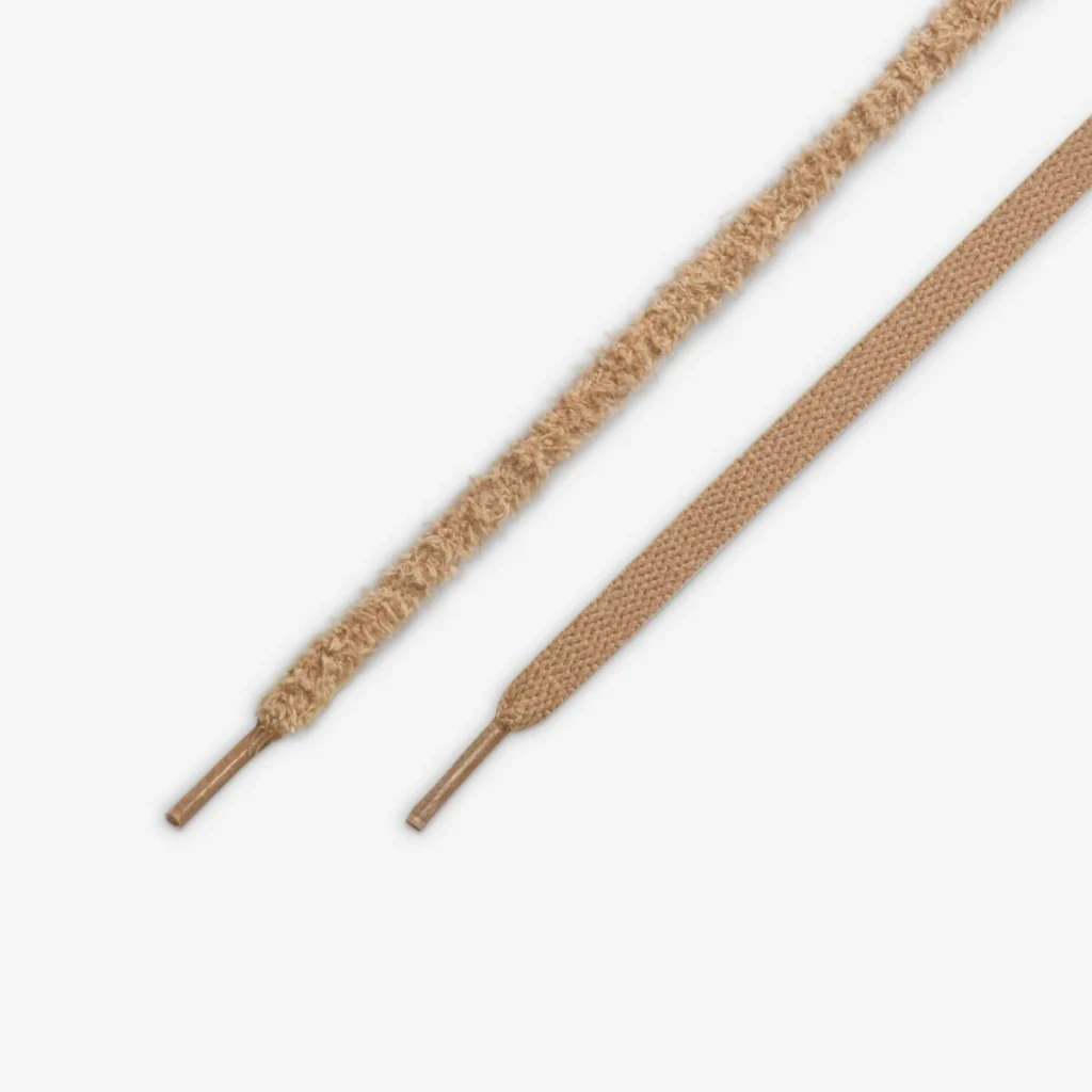 Close-up of two Air Jordan 4 WMNS “Cozy Girl” laces—one fuzzy and textured, the other flat and woven—both in a warm beige tone with matching plastic aglets, displayed on a white background.